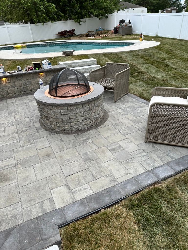 Patio with fire pit, seating, pool in background. Gray pavers, green grass, neutral tones.