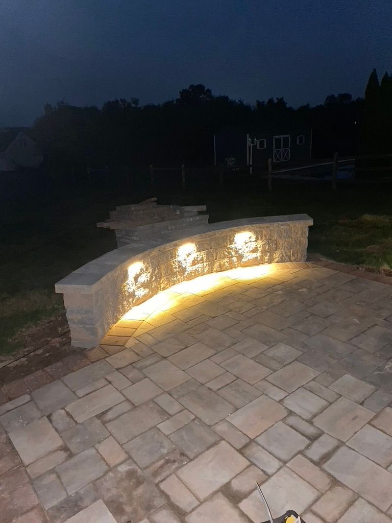 Stone wall with integrated lights curves along a patio at dusk, with trees in the background.