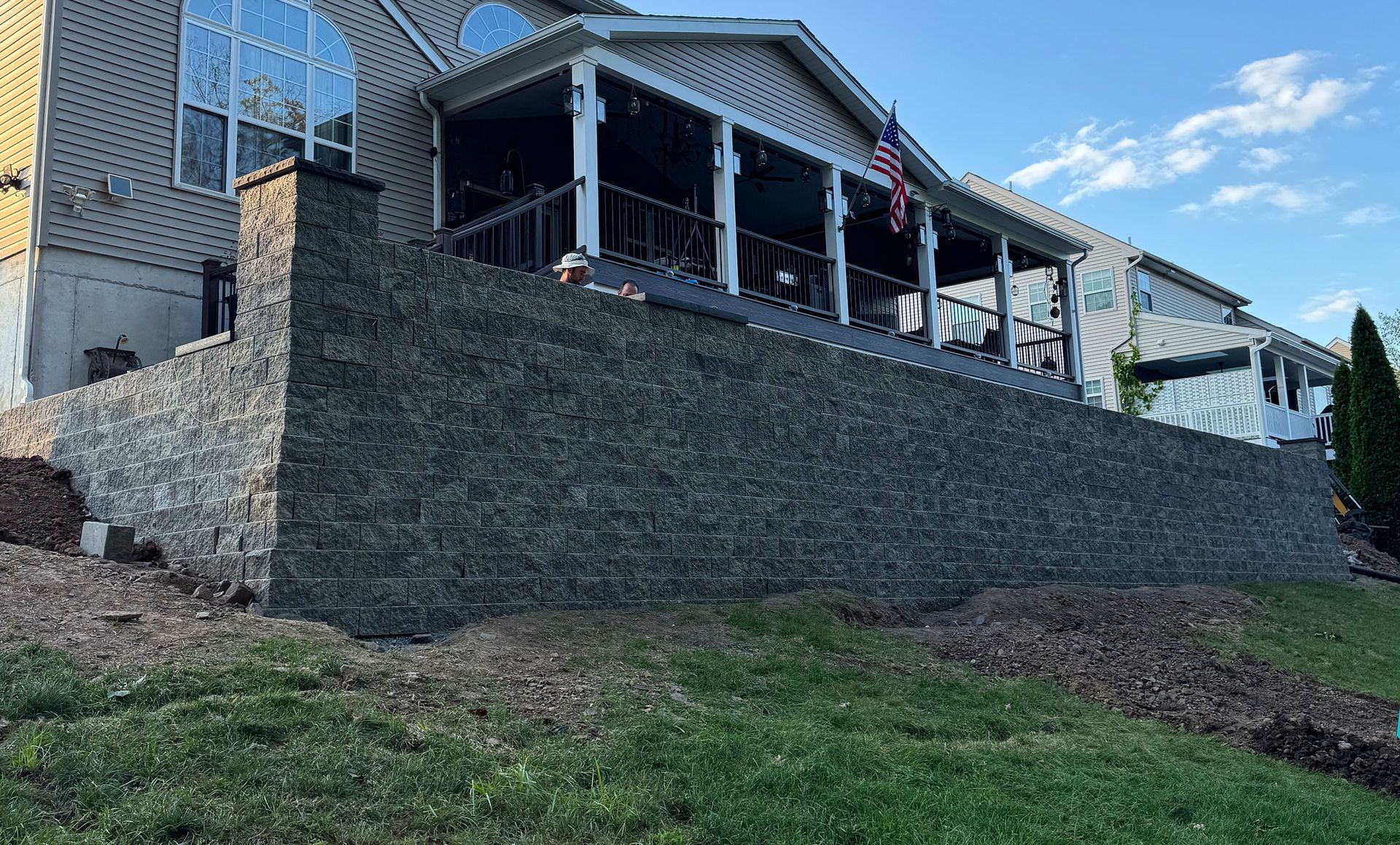 Stone retaining wall in front of a house, supporting a grassy hillside, a porch is visible.