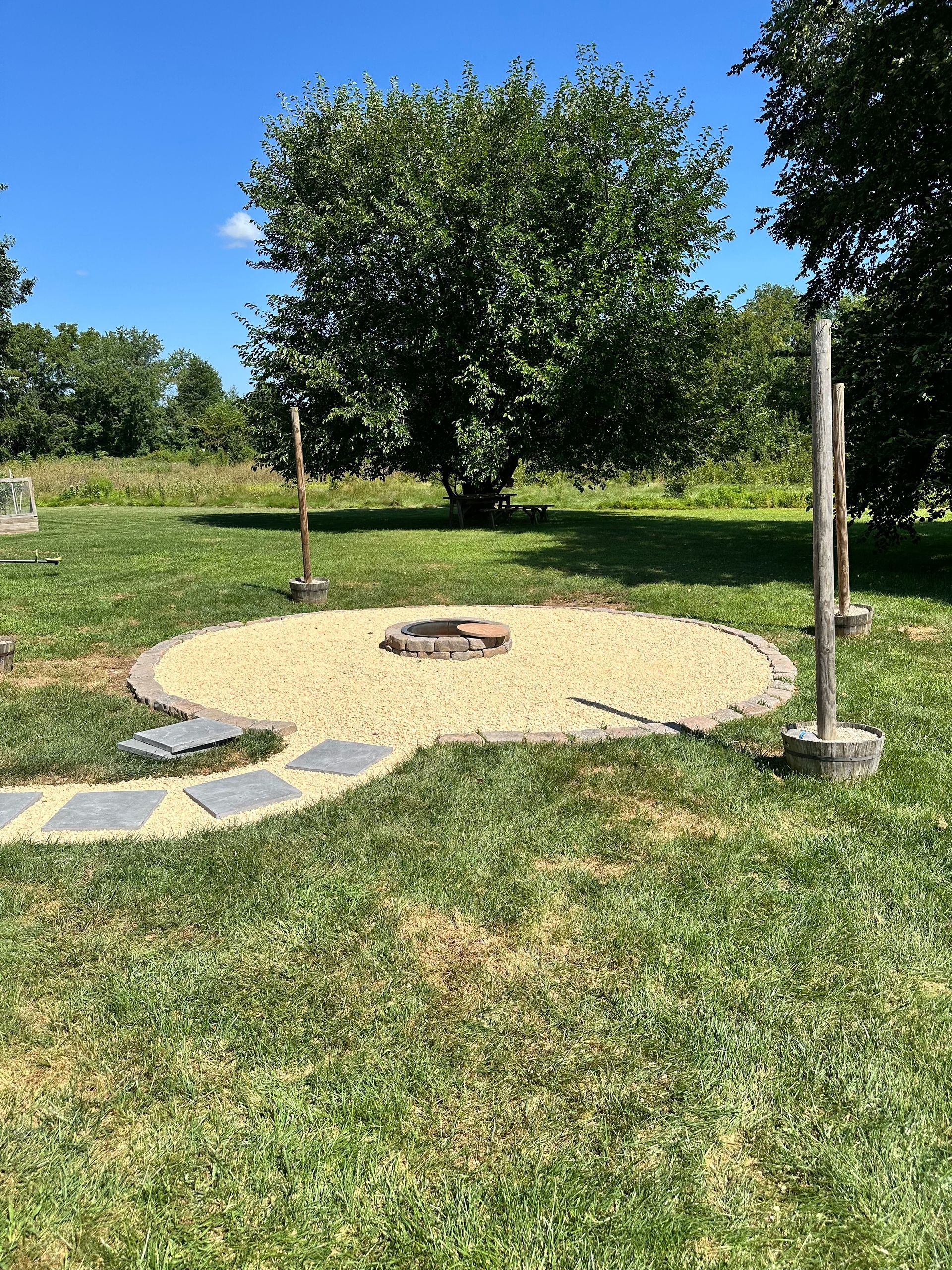 Circular gravel fire pit with stepping stones, wooden posts, and a tree in a grassy area under a blue sky.