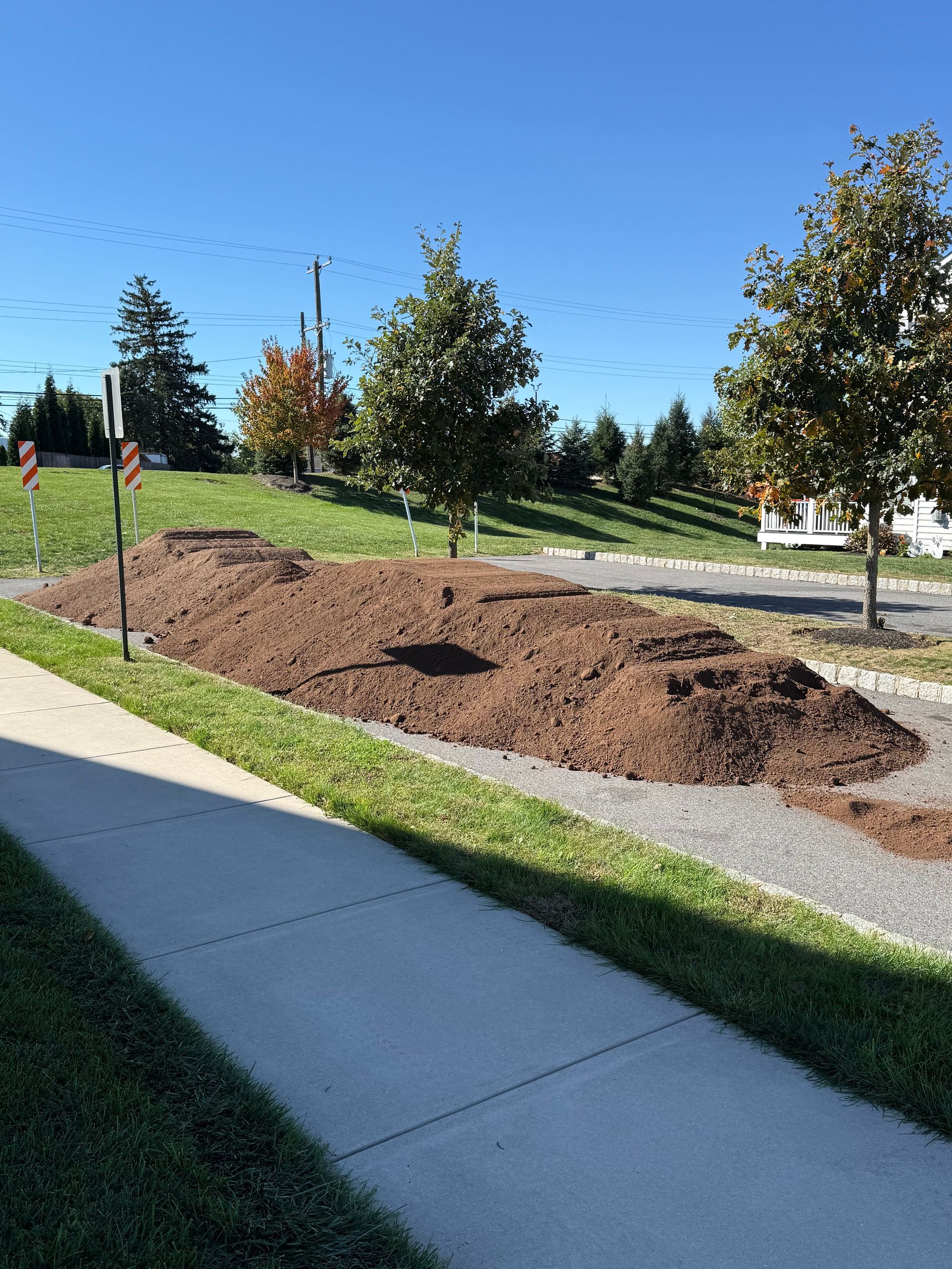 Pile of brown mulch in a landscaped area next to a sidewalk and green grass, trees in the background, blue sky.