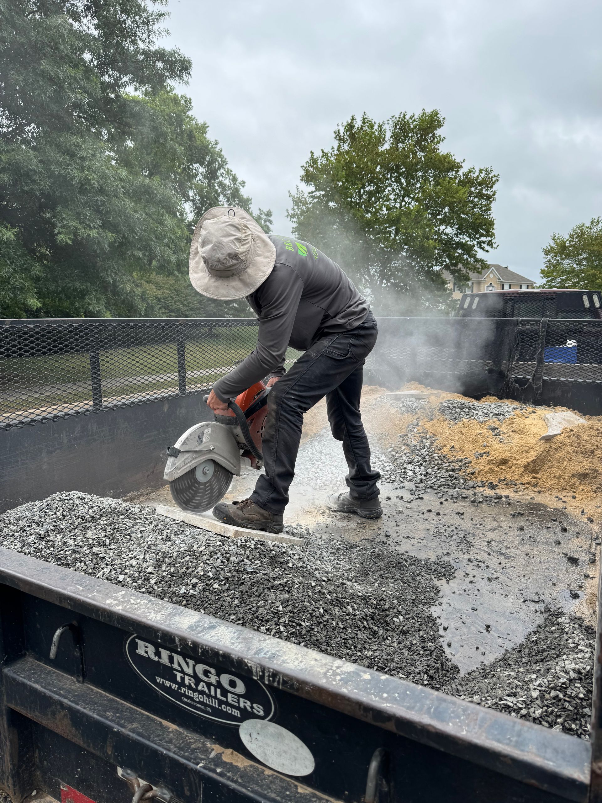 Person cutting gravel with a circular saw in the bed of a dump trailer, creating dust. Gray sky.