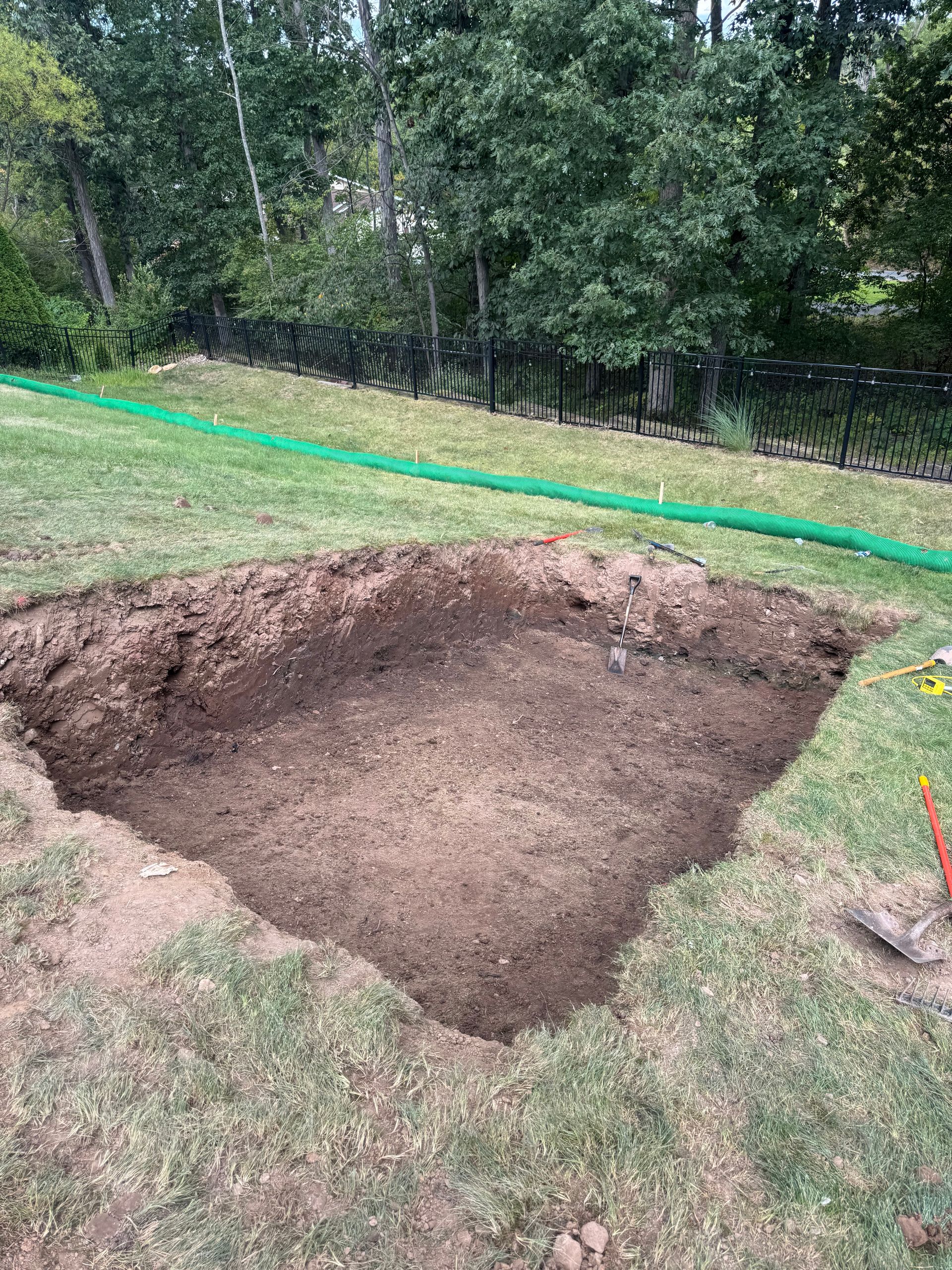 A square excavation pit in a grassy yard, surrounded by green grass and trees.