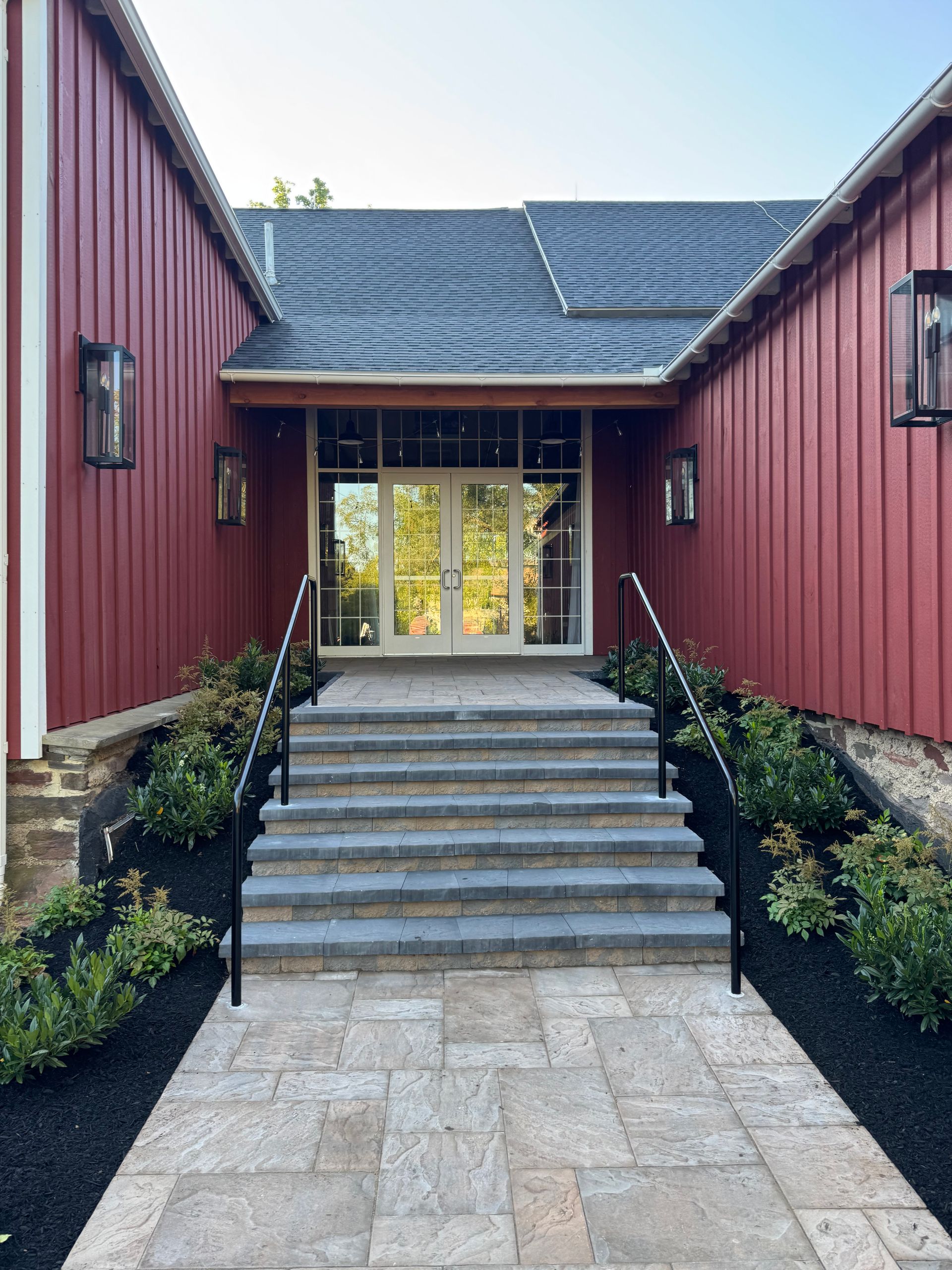 Red-sided building entrance with steps, double doors, and black handrails. Paver path leads to the entry.