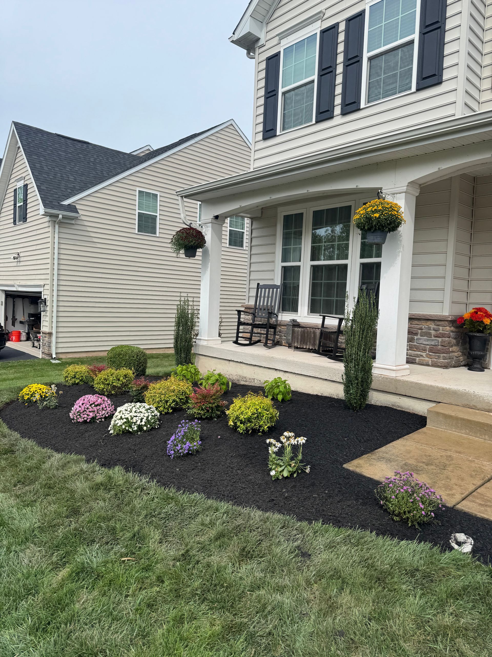 A house with a landscaped front yard featuring colorful flowers and black mulch. Hanging flower baskets on the porch.