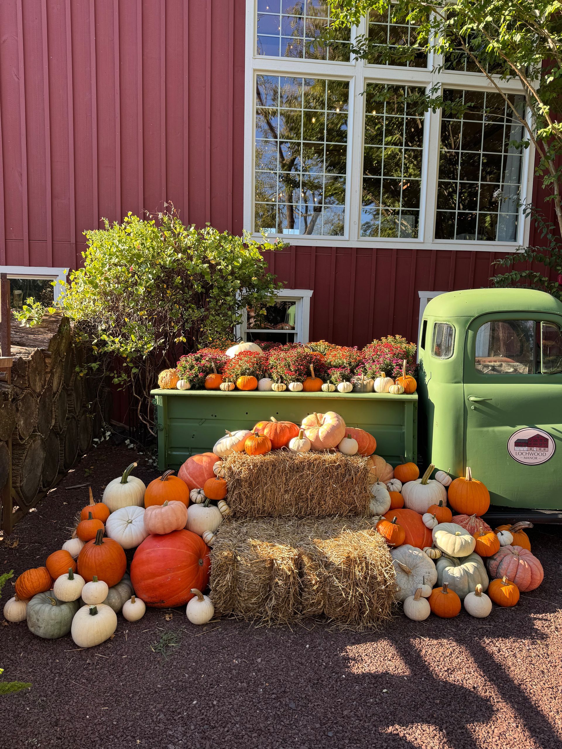Pumpkins and hay bales arranged in front of a green truck and red building. Fall decorations.
