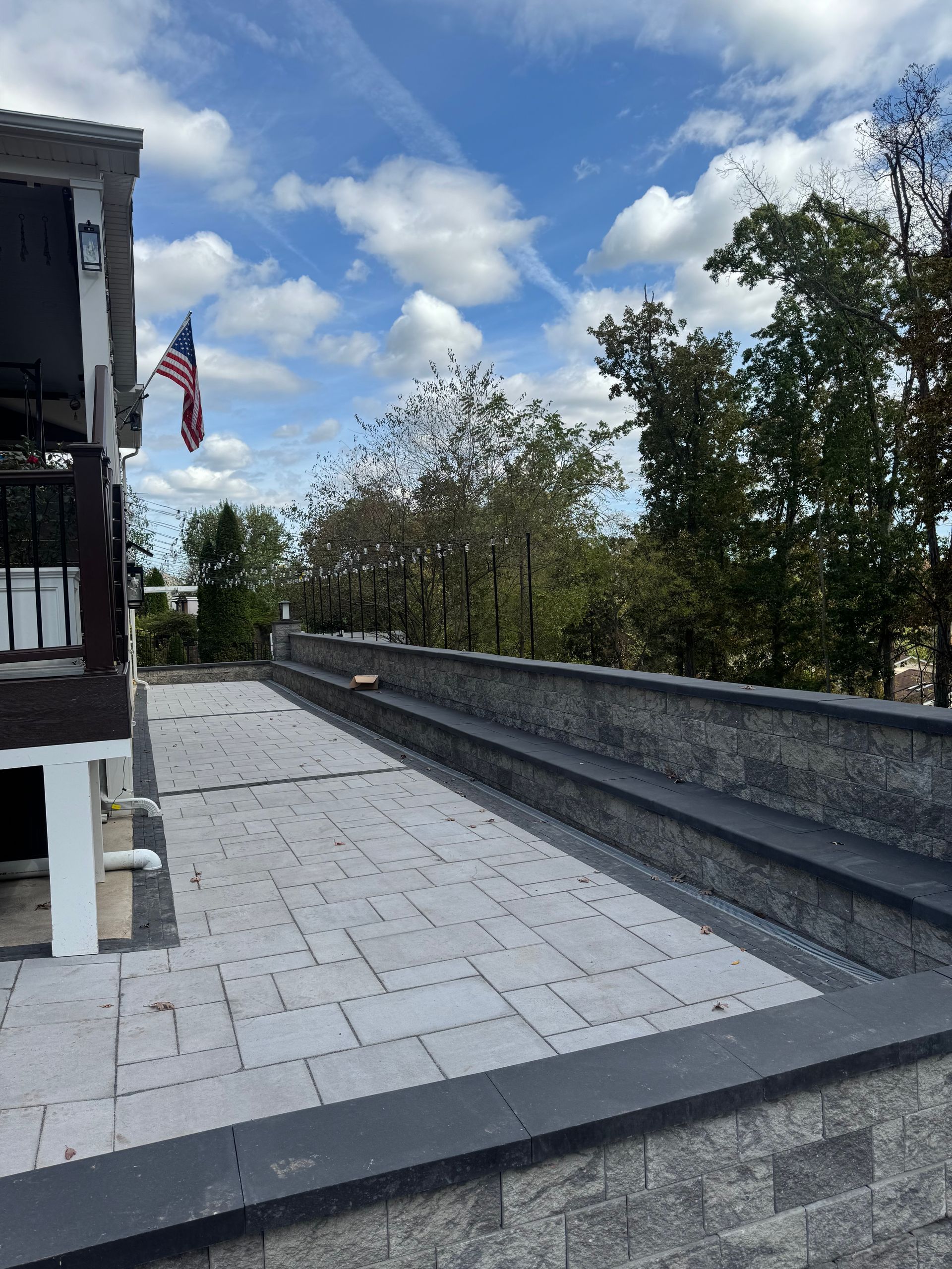 A stone patio with a low wall, an American flag, and trees under a blue sky with clouds.