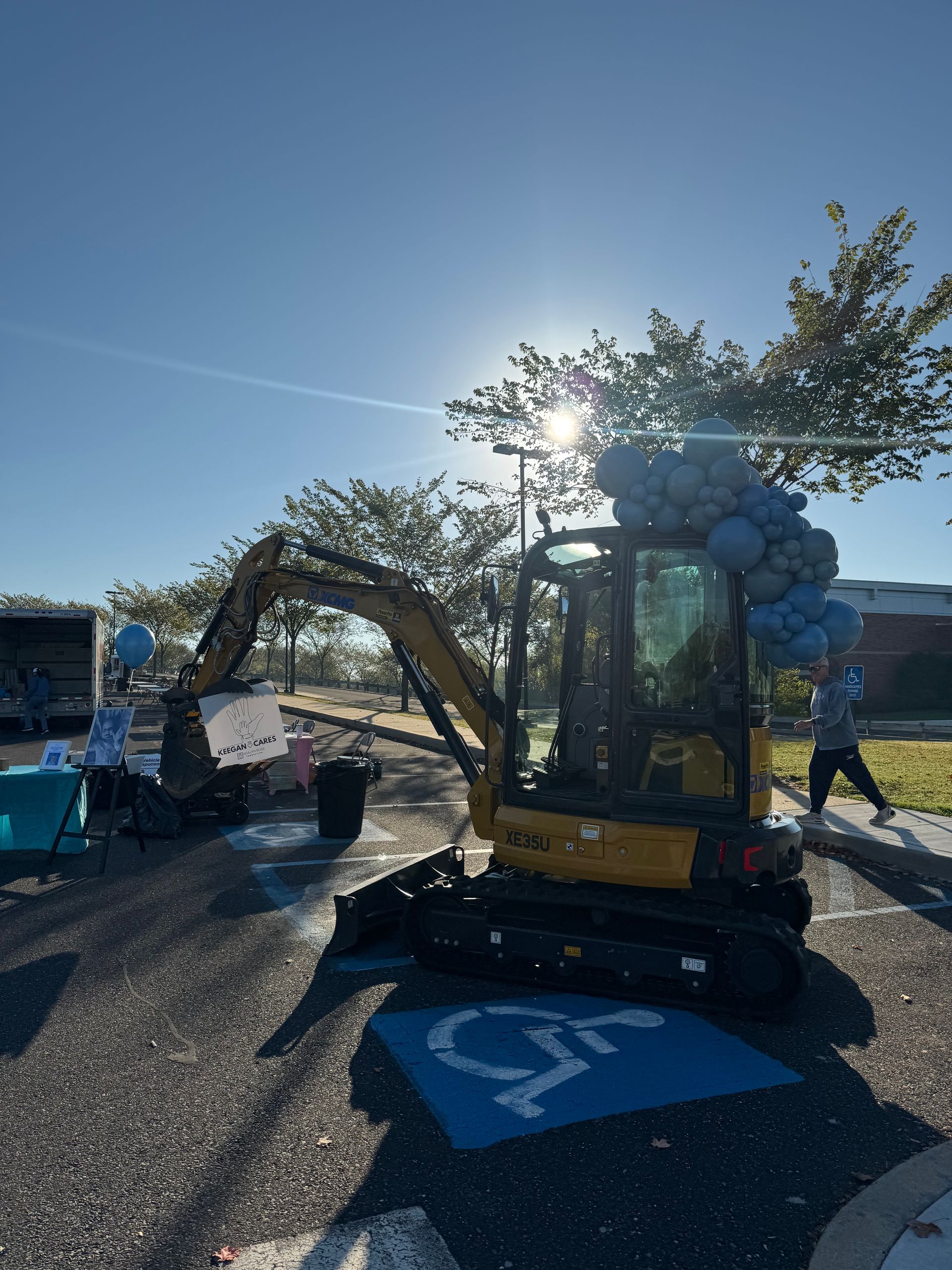 Yellow excavator with balloons in a handicap parking space. A person walks nearby under a sunny sky.