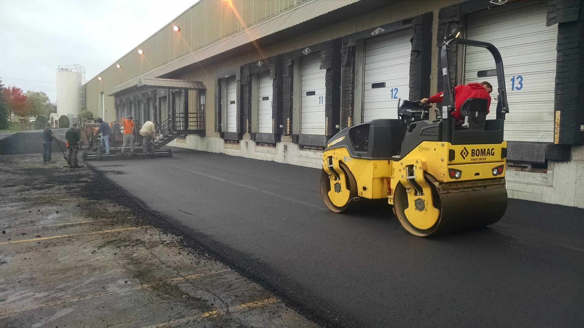 A yellow road roller compacts fresh asphalt near a loading dock, with workers and equipment nearby.