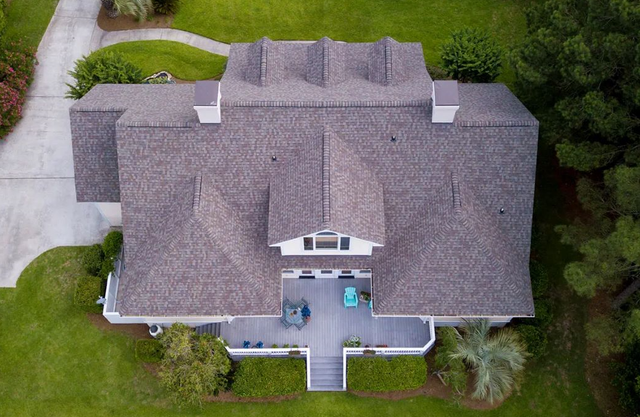 Overhead view of a house with a gray roof and a wooden deck surrounded by green grass and trees.
