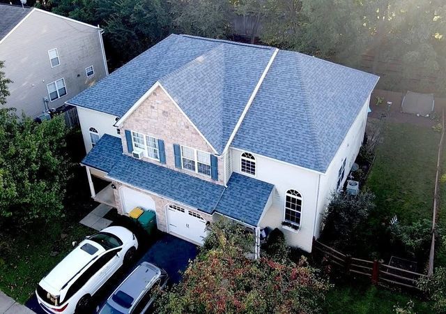 Blue-roofed, two-story house with white siding and a driveway with two cars. Trees and a fence surround it.