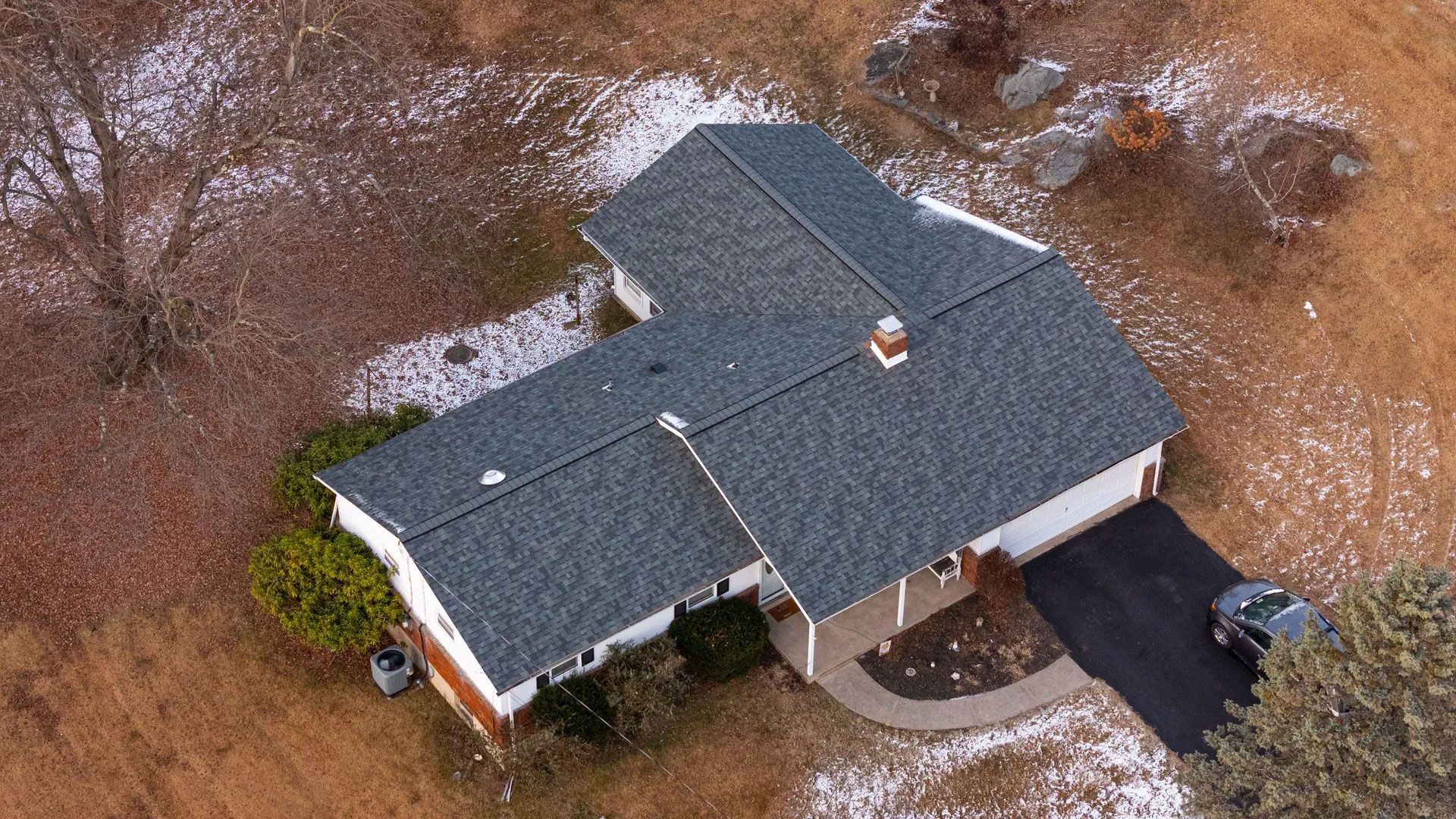 Overhead view of a house with a dark gray roof, black driveway, and surrounding brown and snowy landscape.