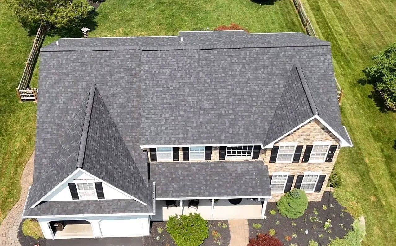 Aerial view of a gray-roofed house with black shutters, surrounded by green grass and a wooden fence.