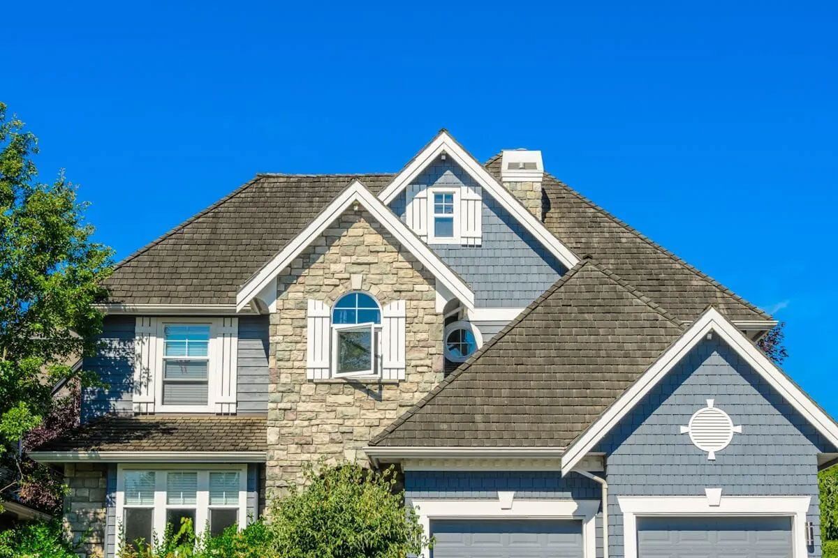 Two-story house with gray shingles, stone and blue siding, white trim, and a clear blue sky.