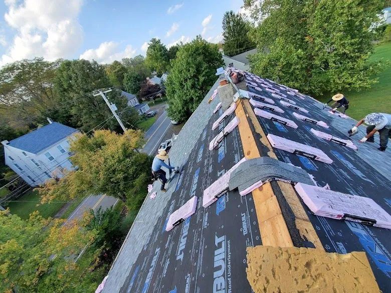 Roofers working on a residential roof, installing underlayment and shingles. Sunny day, surrounded by trees.