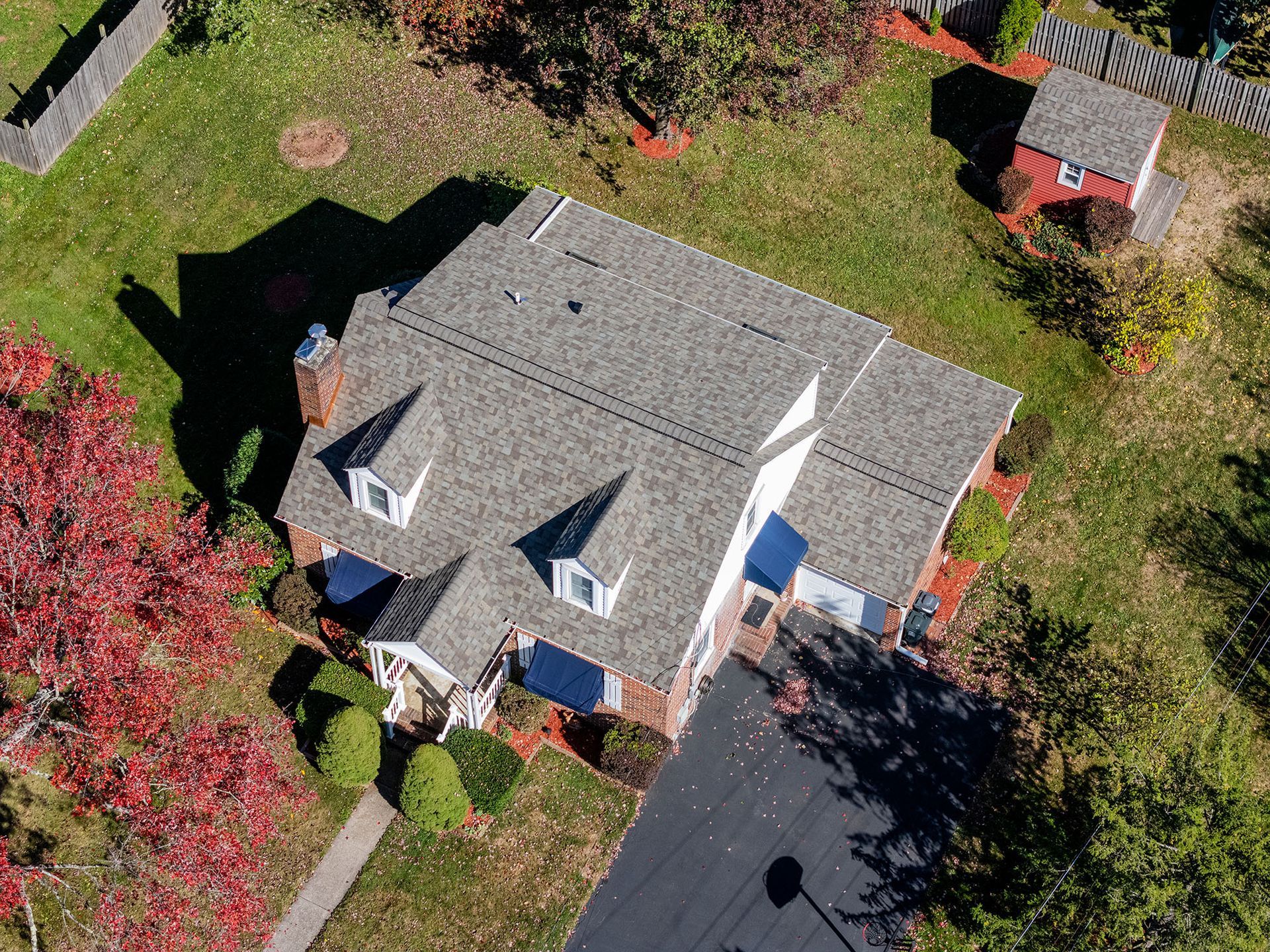 Aerial view of a house with a gray roof and dormers, surrounded by lawn and fall foliage.