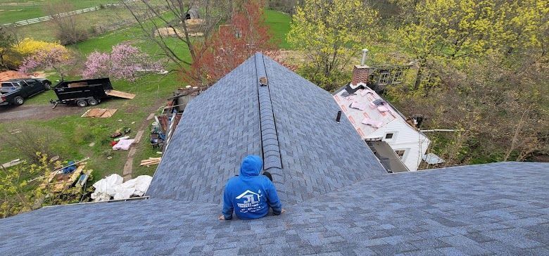 A person in a blue hoodie on a roof, working on shingles. A trailer and trees are in the background.