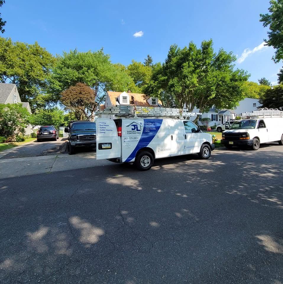 White service van with ladder parked on a street; other vehicles and houses in the background. Blue sky and trees.