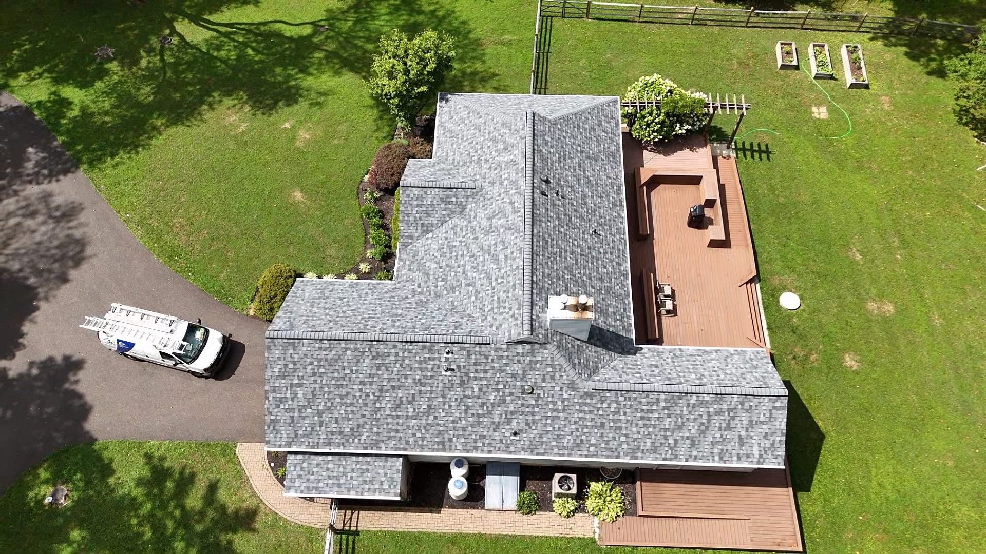 Overhead view of a house with a gray roof, brown deck, and green lawn. A white vehicle is in the driveway.