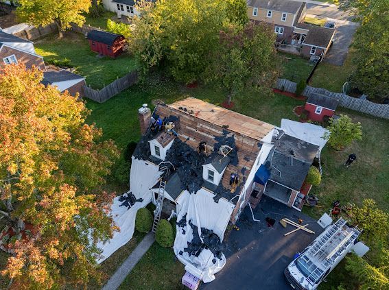 Aerial view of house roof damage covered with tarps; workers on roof; fire truck present.