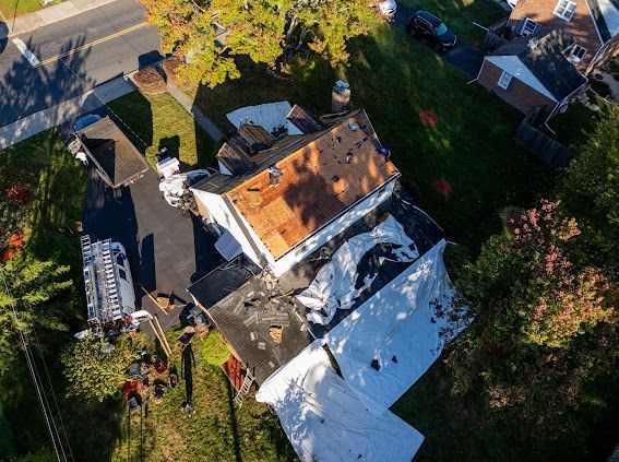 Aerial view of a house with significant roof damage and debris, asphalt driveway, and surrounding green lawn.