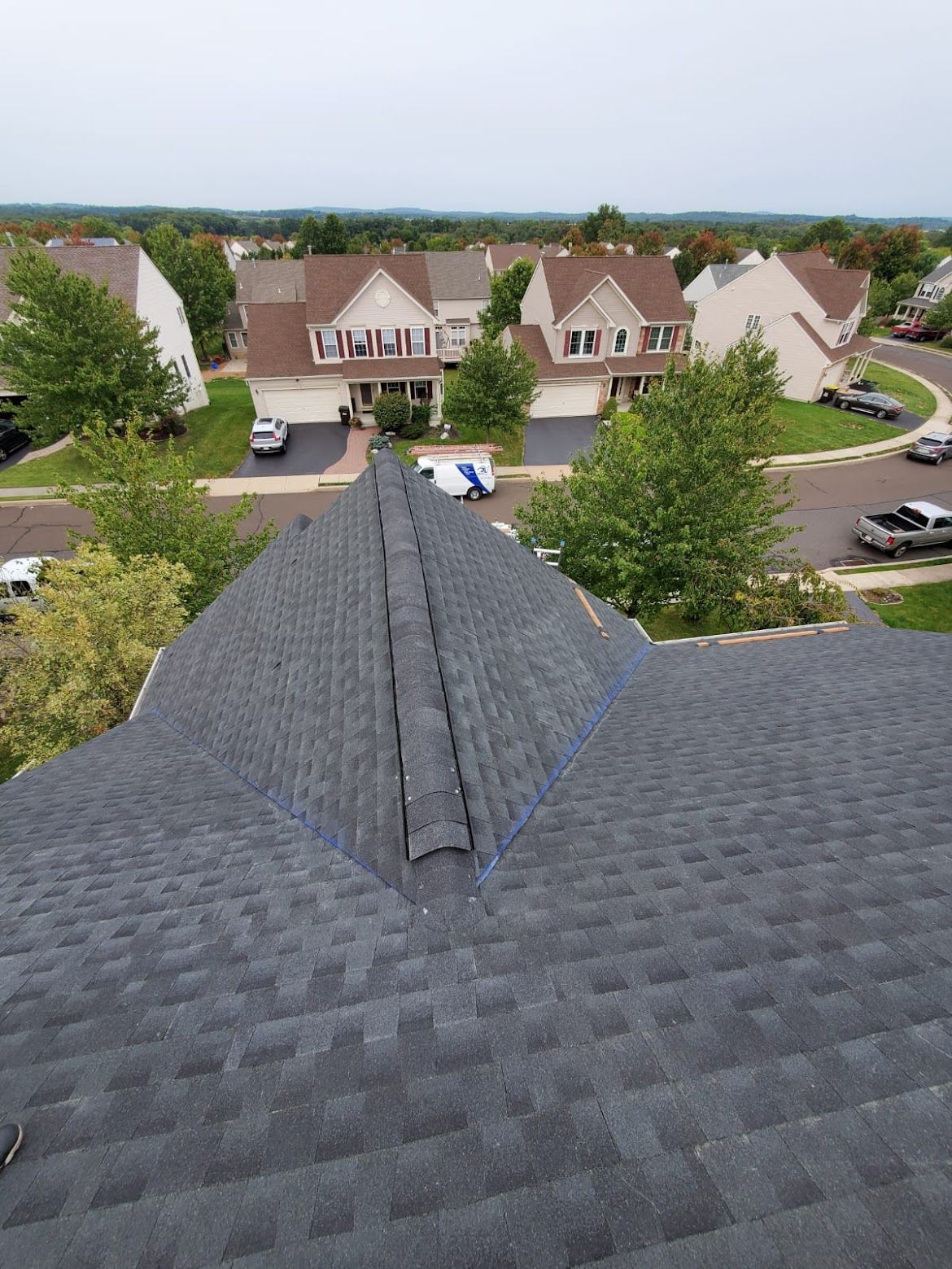 View of a dark gray shingle roof in a suburban neighborhood.  Other houses and trees are visible.