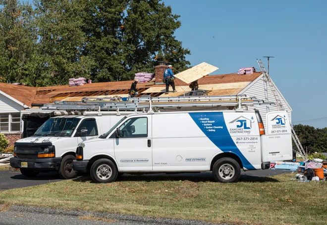 Roofers working on a house roof on a sunny day. White company vans with ladders parked nearby.