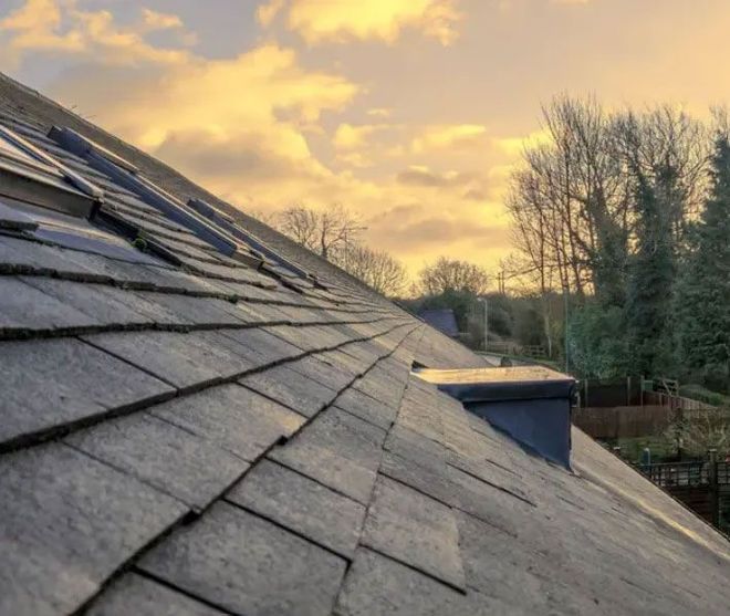 Slate roof on a building with a chimney, trees, and a cloudy sunset sky.