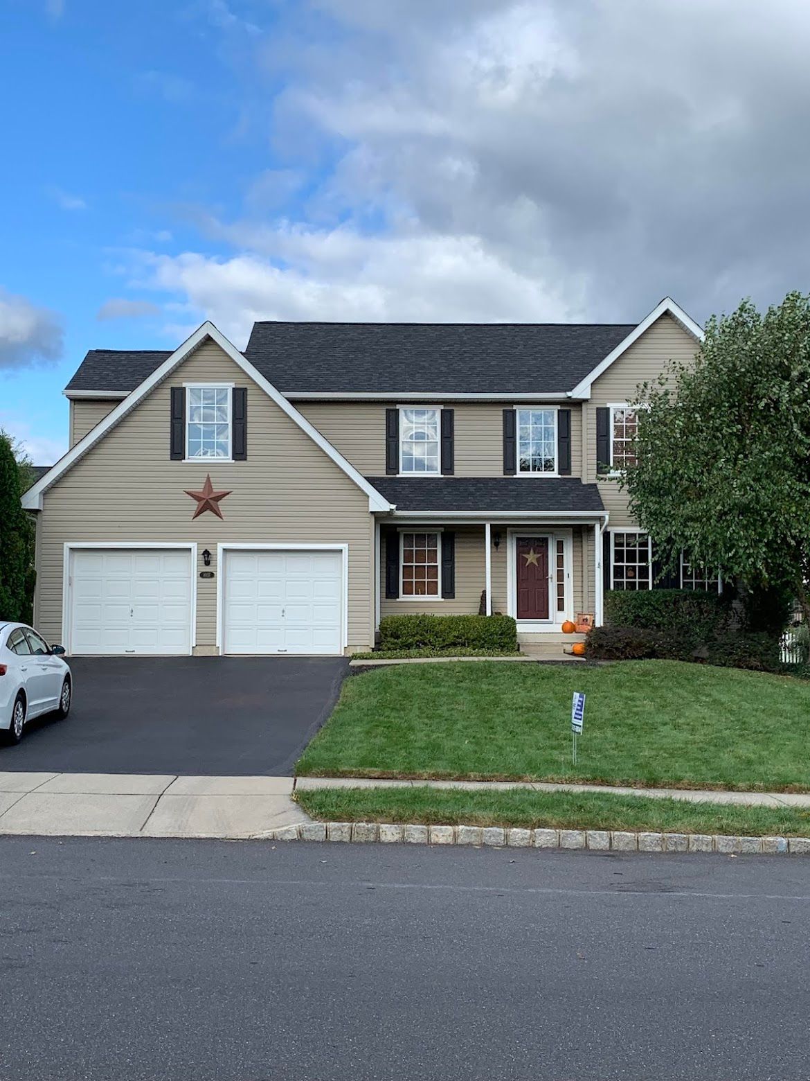Two-story beige house with a black roof, two-car garage, and a green lawn under a cloudy sky.