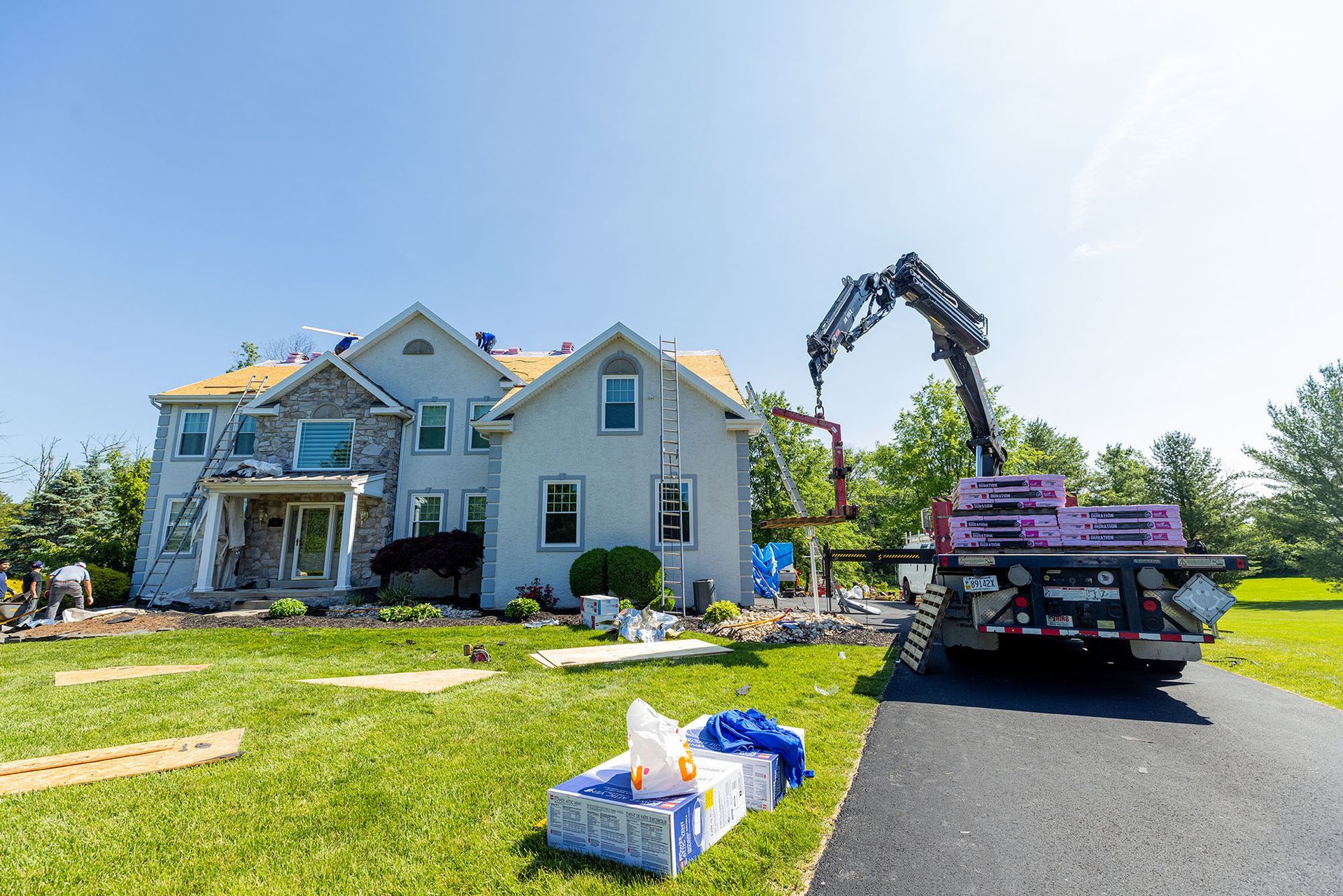 Roofers using a crane to lift roofing materials onto a two-story house on a sunny day.