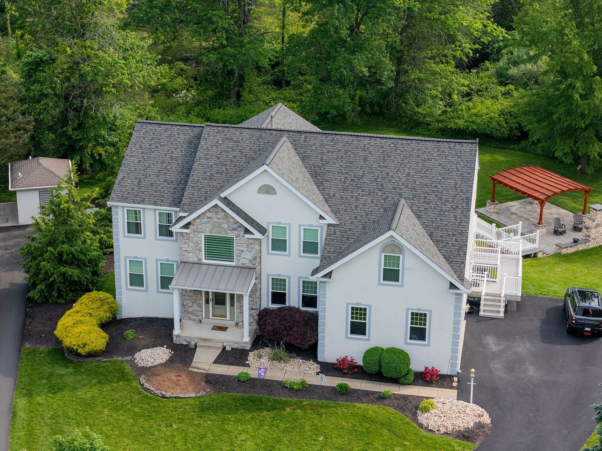 Two-story gray house with stone facade, dark roof, and white trim. Landscaping with shrubs and trees.