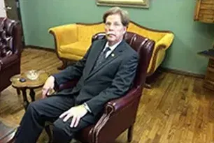 Man seated in a burgundy armchair in a green room with wooden floors and yellow sofa nearby