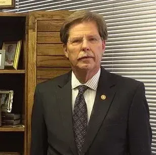 Man in a dark suit and tie standing indoors beside wooden paneling and window blinds