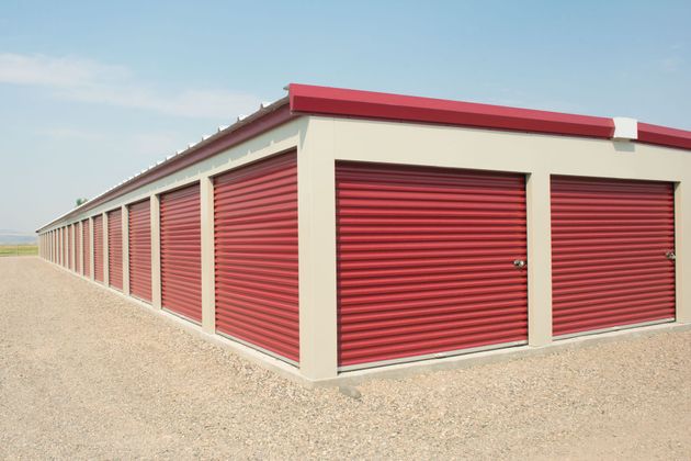 A long, beige self-storage building with red roll-up doors on a gravel lot under a clear sky.