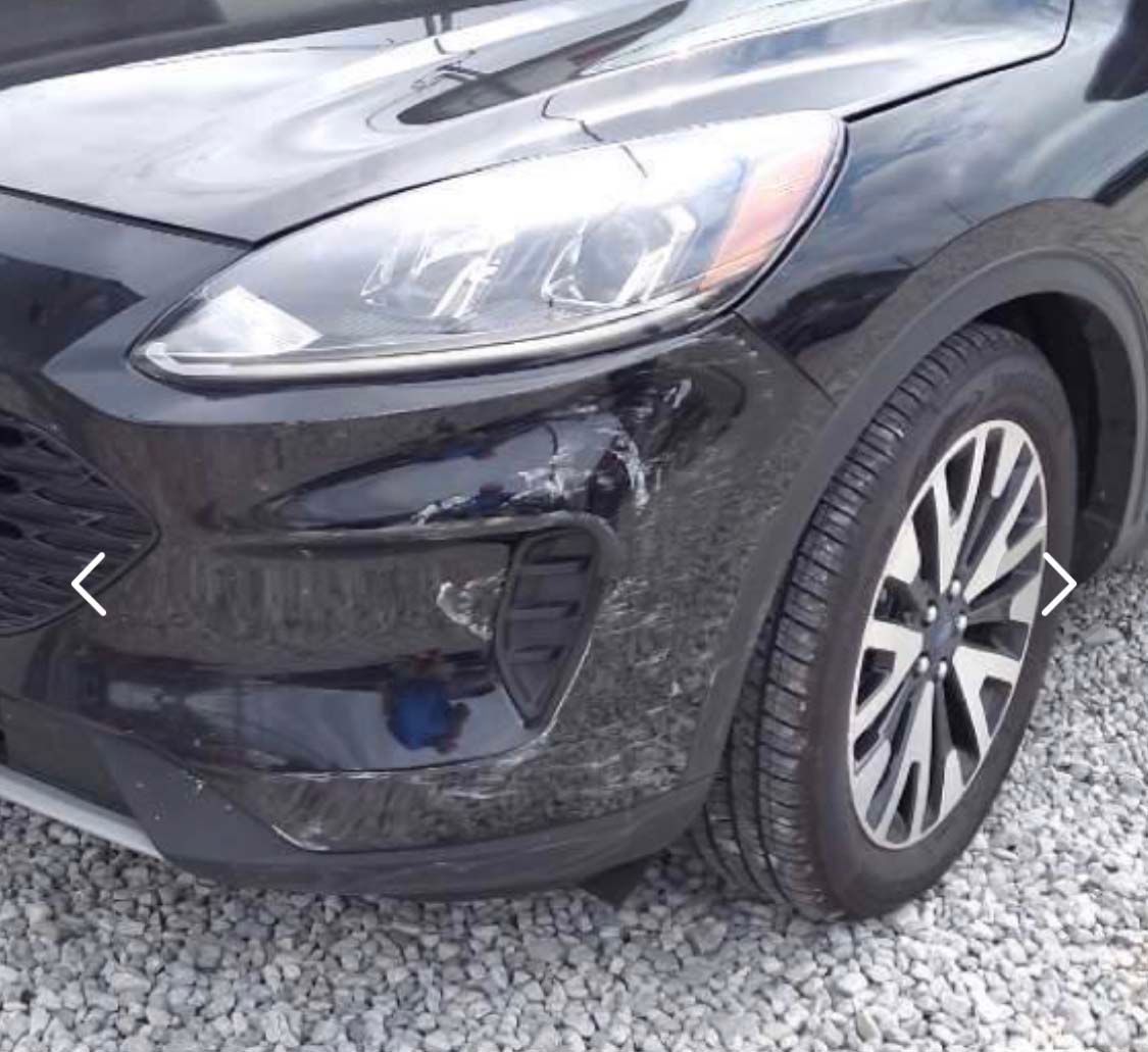 A black car with a damaged bumper is parked on gravel.