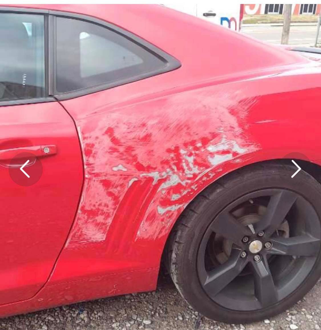 A red car with a damaged side is parked on gravel