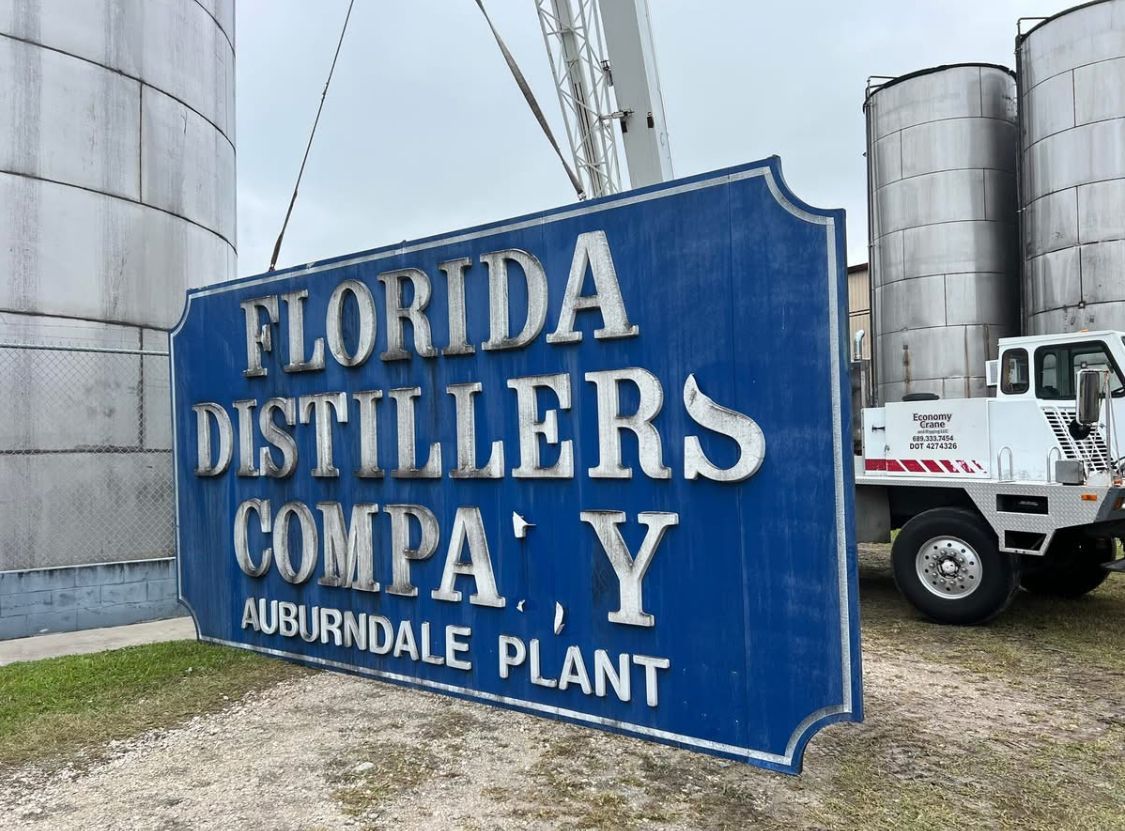 Sign for Florida Distillers Company, Auburndale Plant. Blue sign with silver lettering against a backdrop of tanks and a white truck.