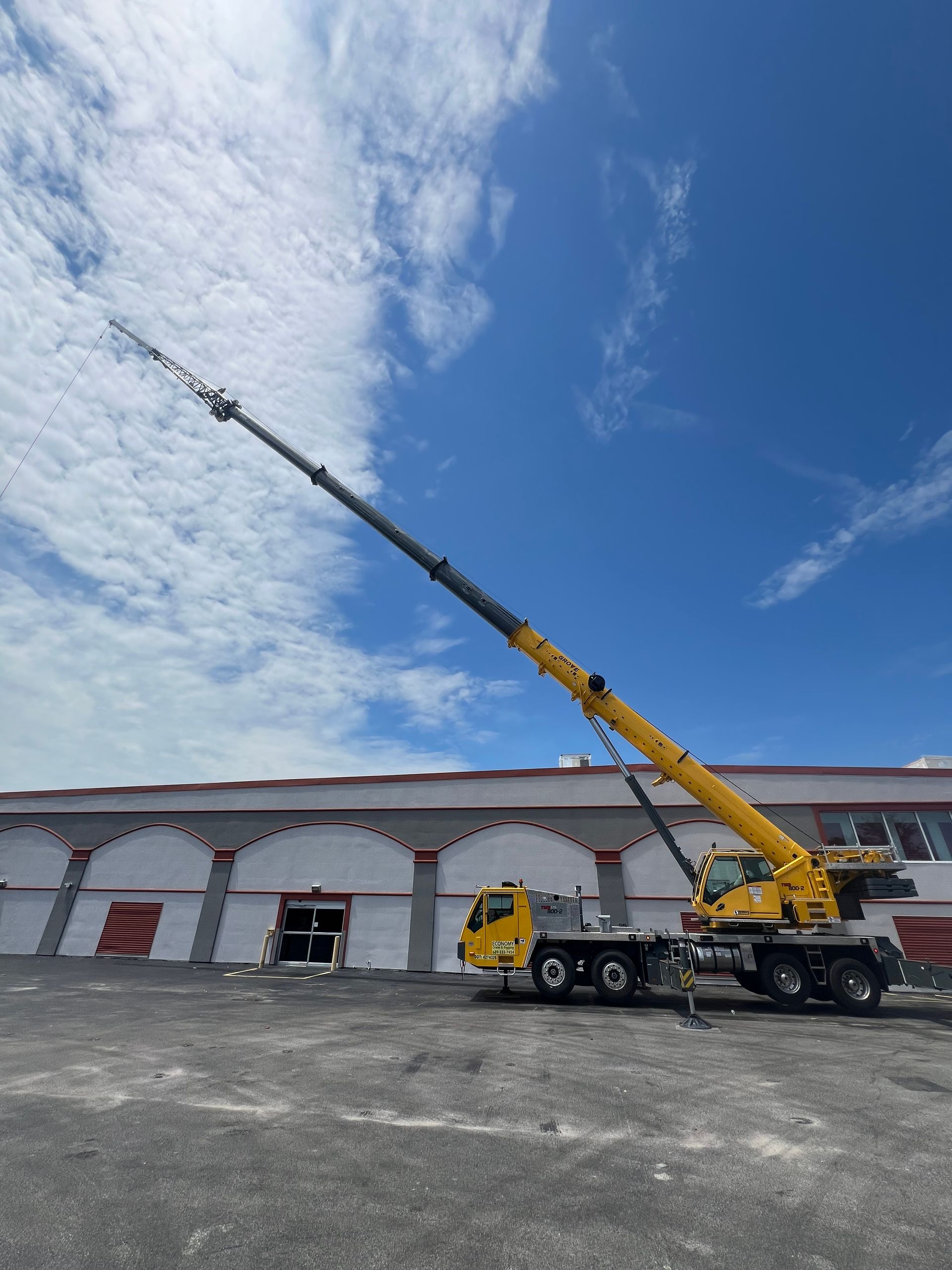 Yellow crane extending its arm skyward near a warehouse under a bright blue sky with clouds.