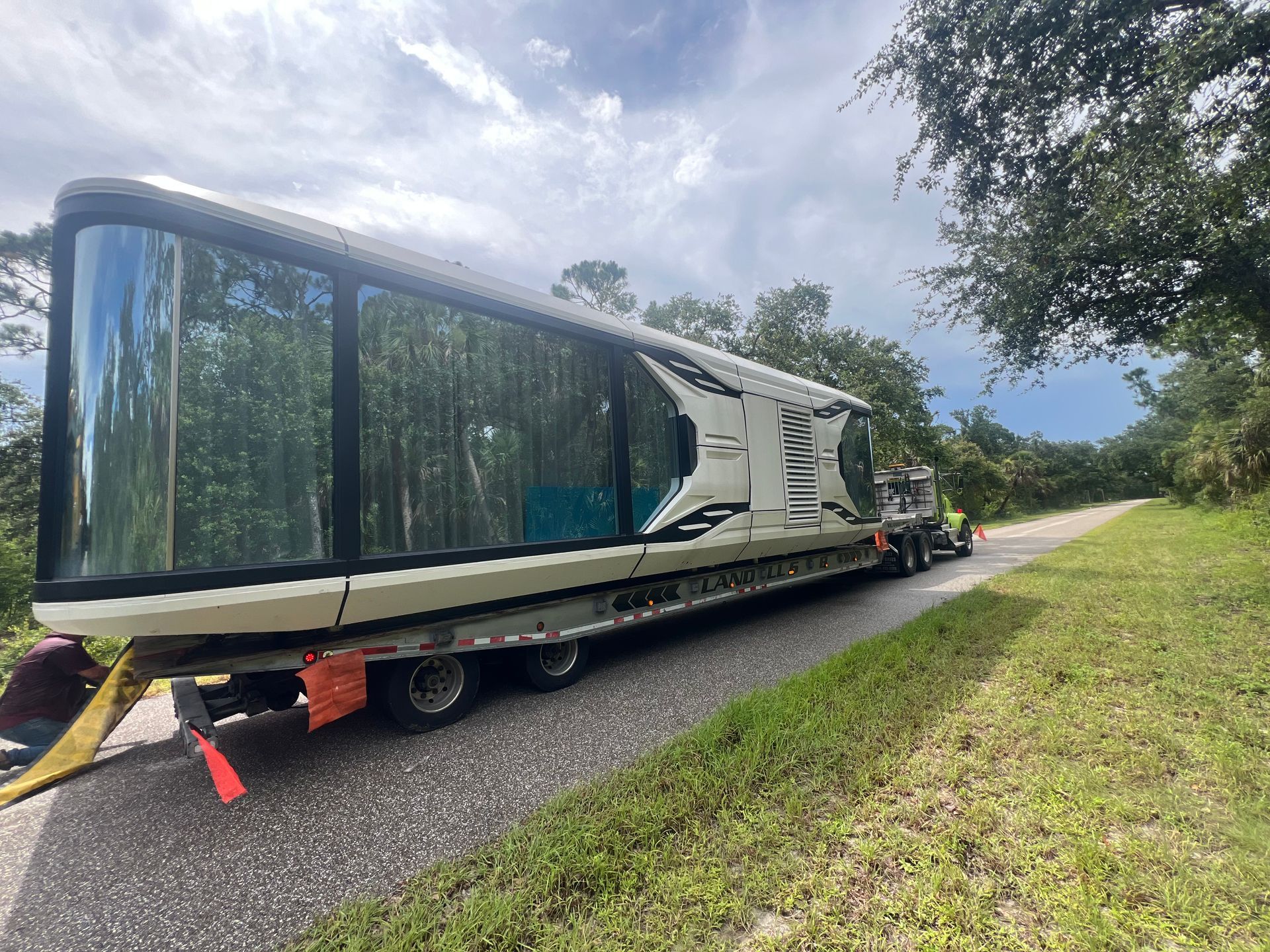 A long, modern trailer home is being transported on a truck on a road lined with trees.