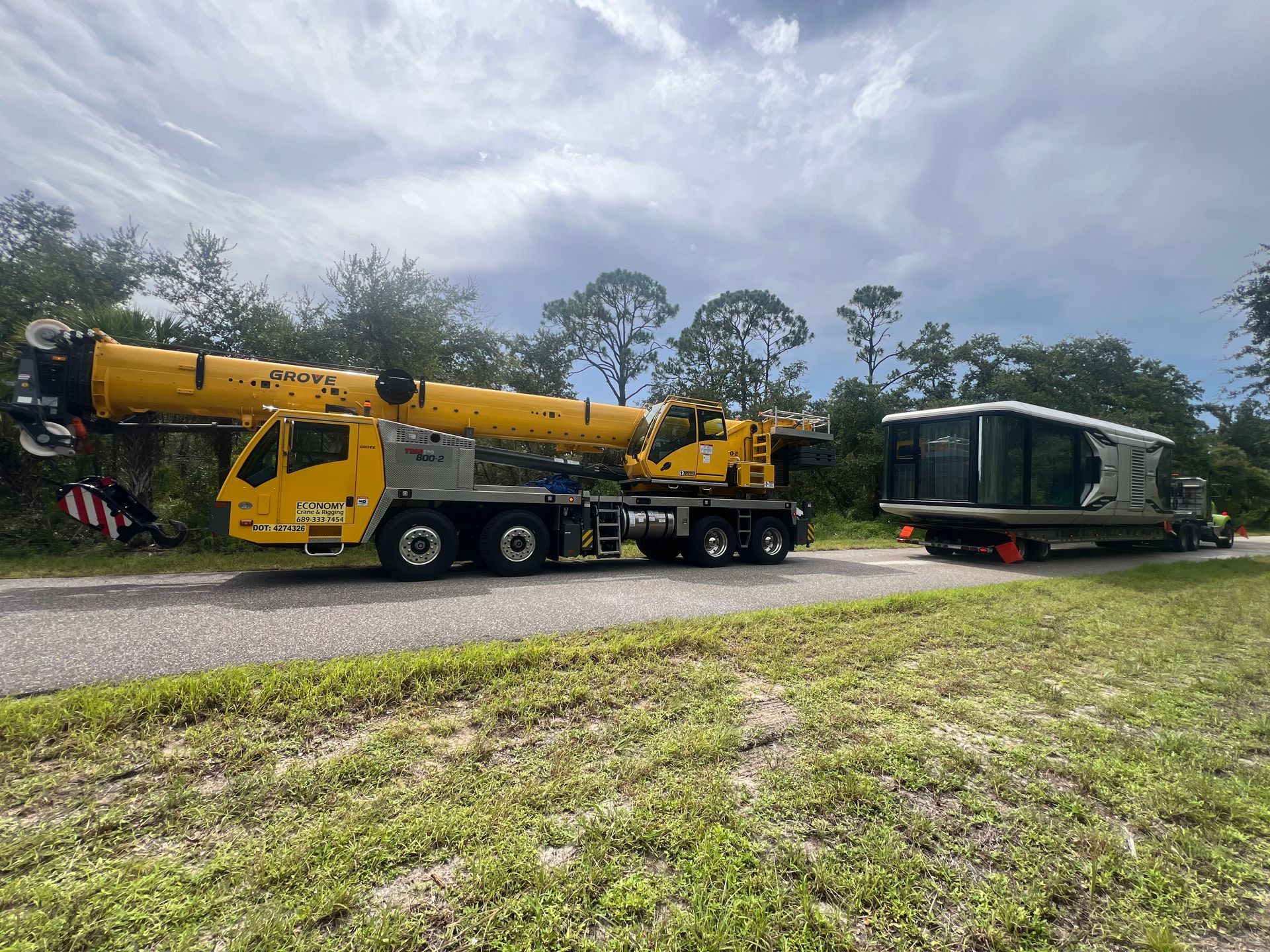 Yellow crane next to a small building on a trailer on a road in a wooded area.