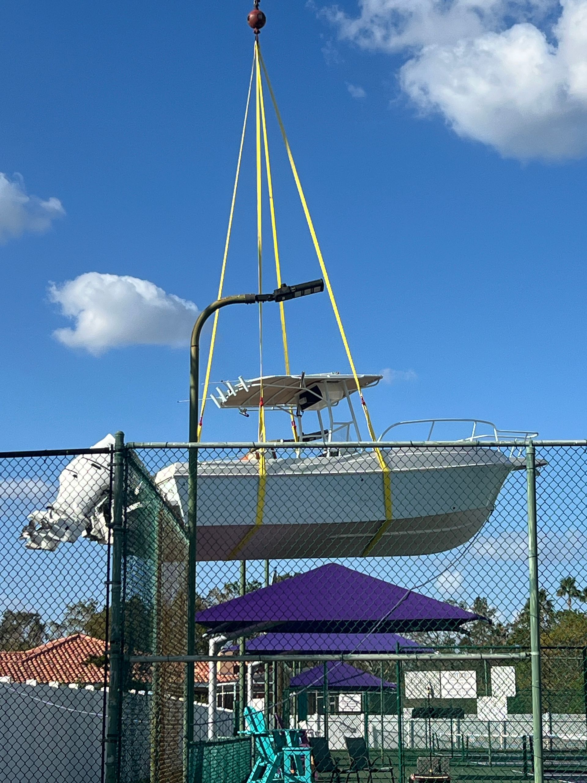 A boat suspended by yellow straps, lifted by a crane over a chain-link fence with purple umbrellas visible below.