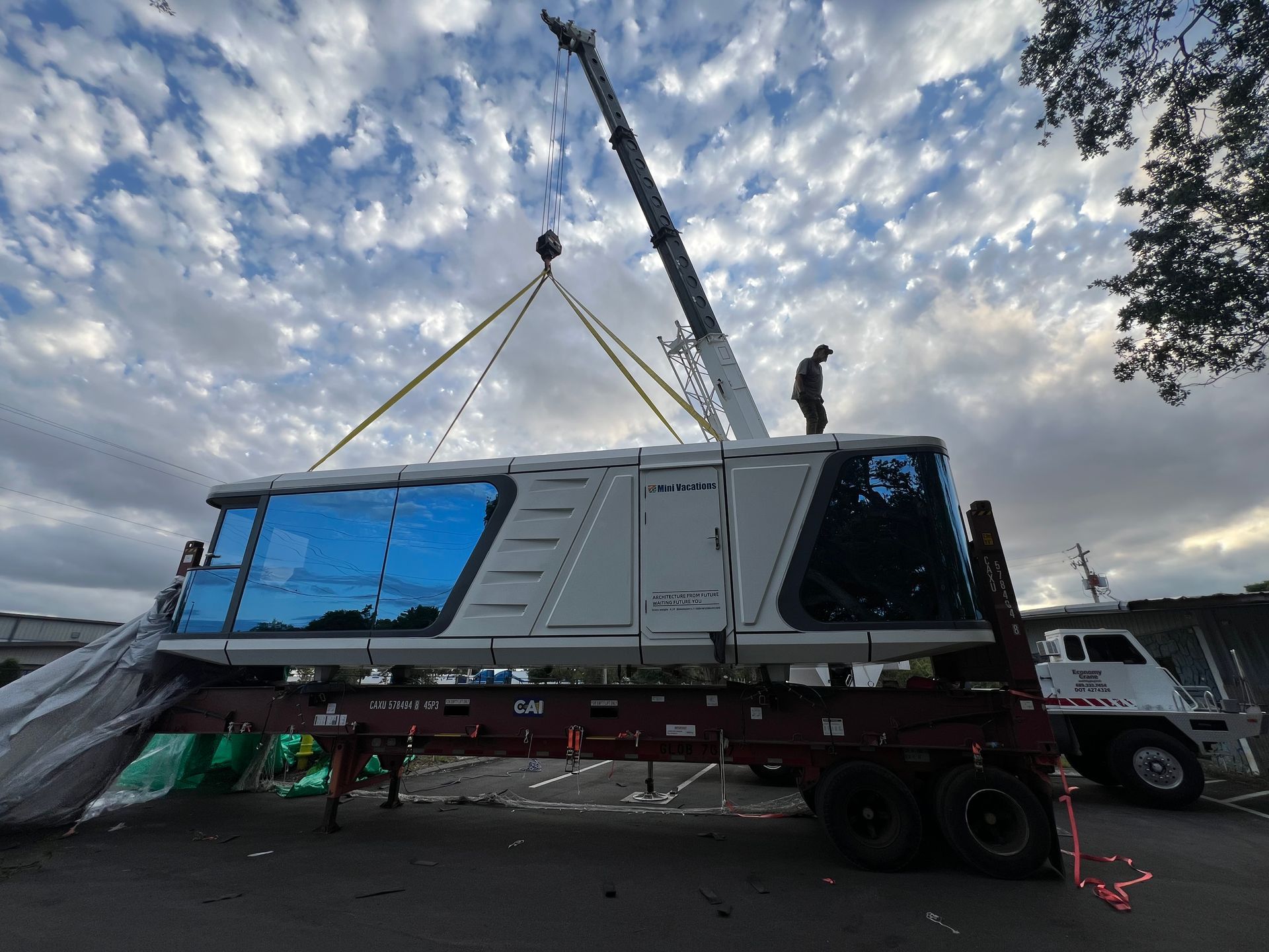 A crane lifting a white rail car from a flatbed truck under a cloudy sky.