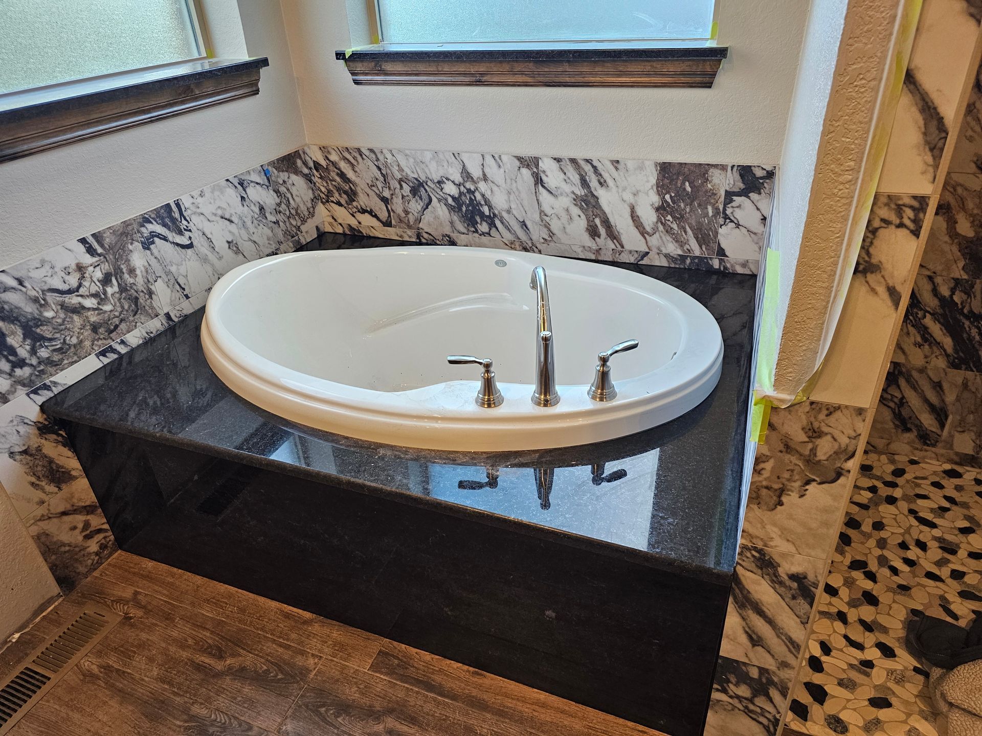 Oval bathtub set in dark granite surround, brown tile floor, and window trim with a marble backsplash.