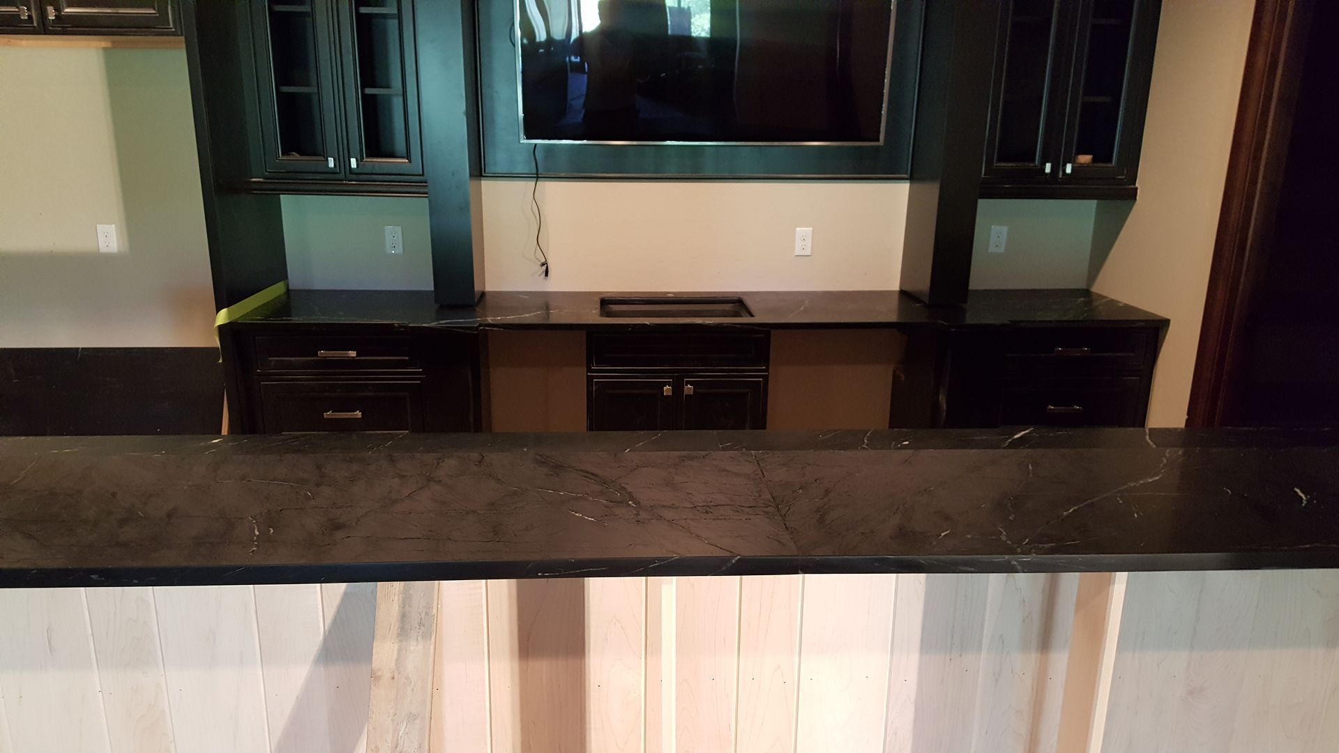 Dark cabinets, countertop and TV against a beige wall, above a white-paneled bar.