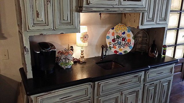 Distressed white cabinets above a countertop with a sink, coffee maker, and colorful decorations.