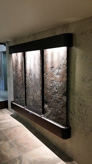 Wall-mounted water feature with three textured panels, lit from above, in a dark-colored frame, set on tiled floor.