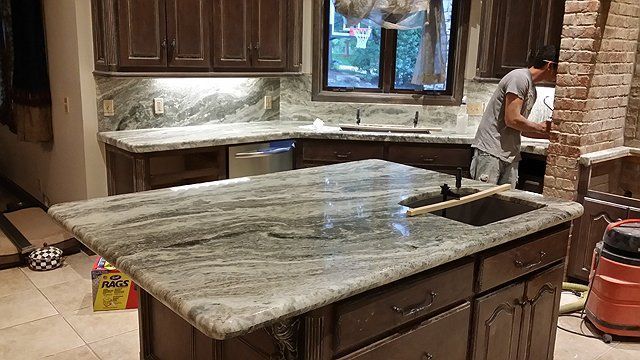 Kitchen with granite countertops and dark cabinets; a person works near a brick column.