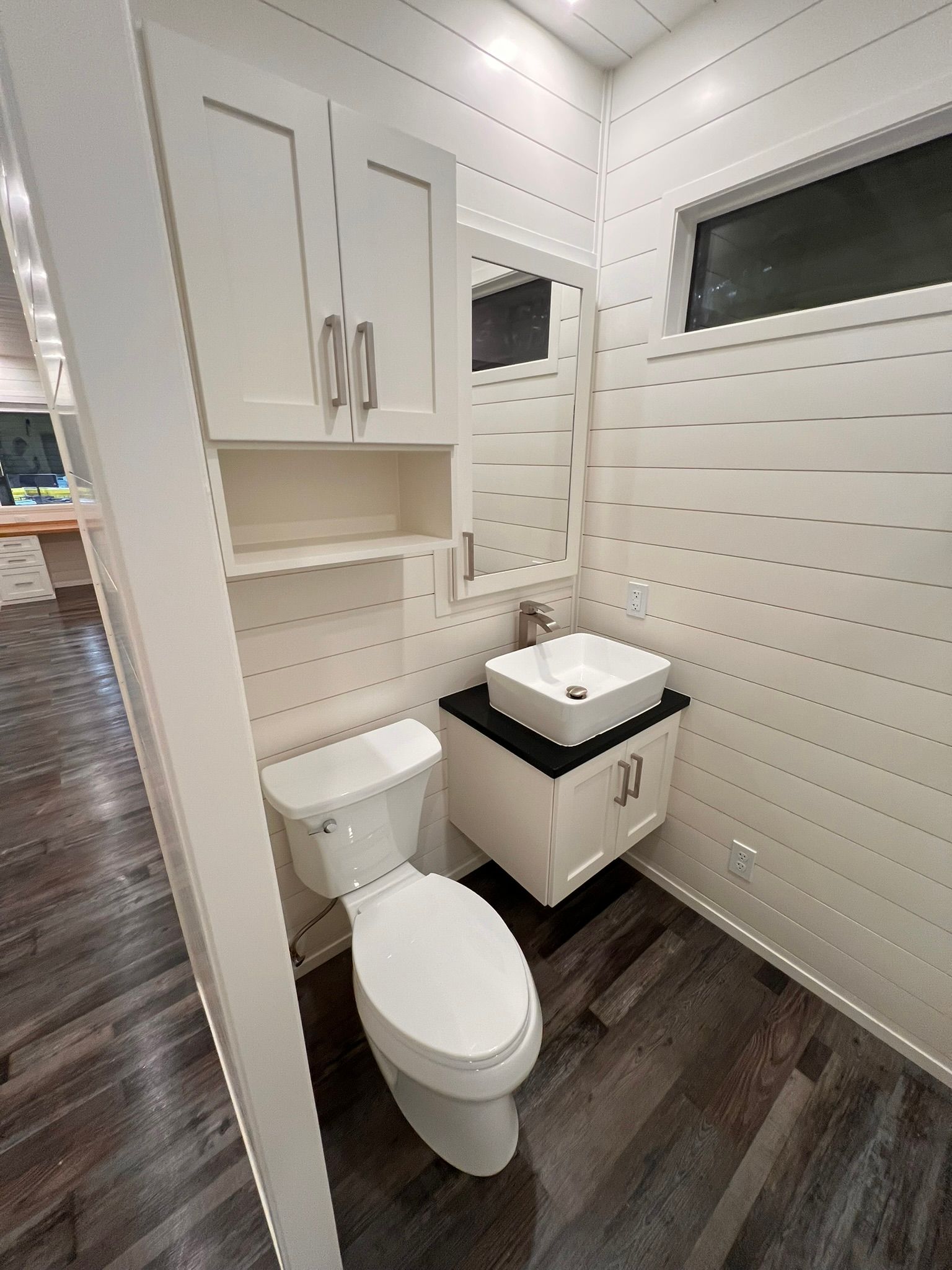 Small bathroom with white shiplap walls, toilet, vanity, and cabinet; dark wood-look floor.
