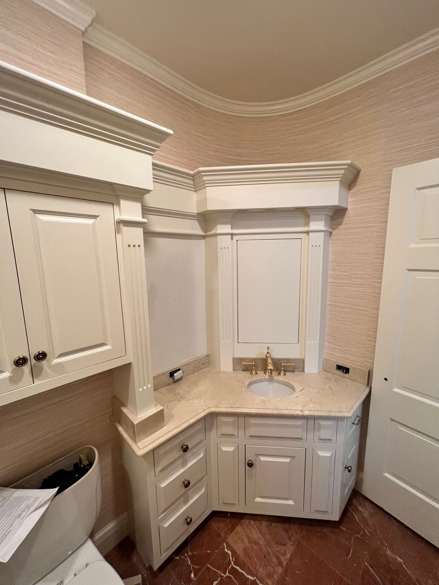 Corner bathroom vanity with white cabinets, gold fixtures, beige countertop, and decorative trim.