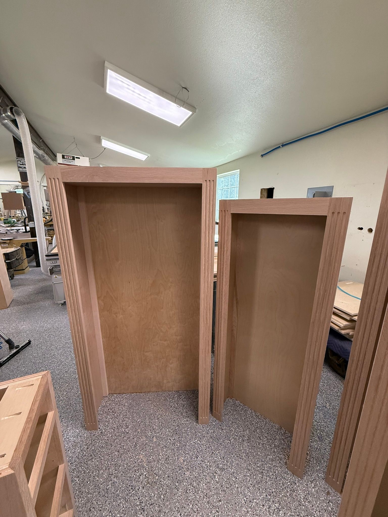 Two unfinished wooden door frames stand on a speckled floor in a workshop.